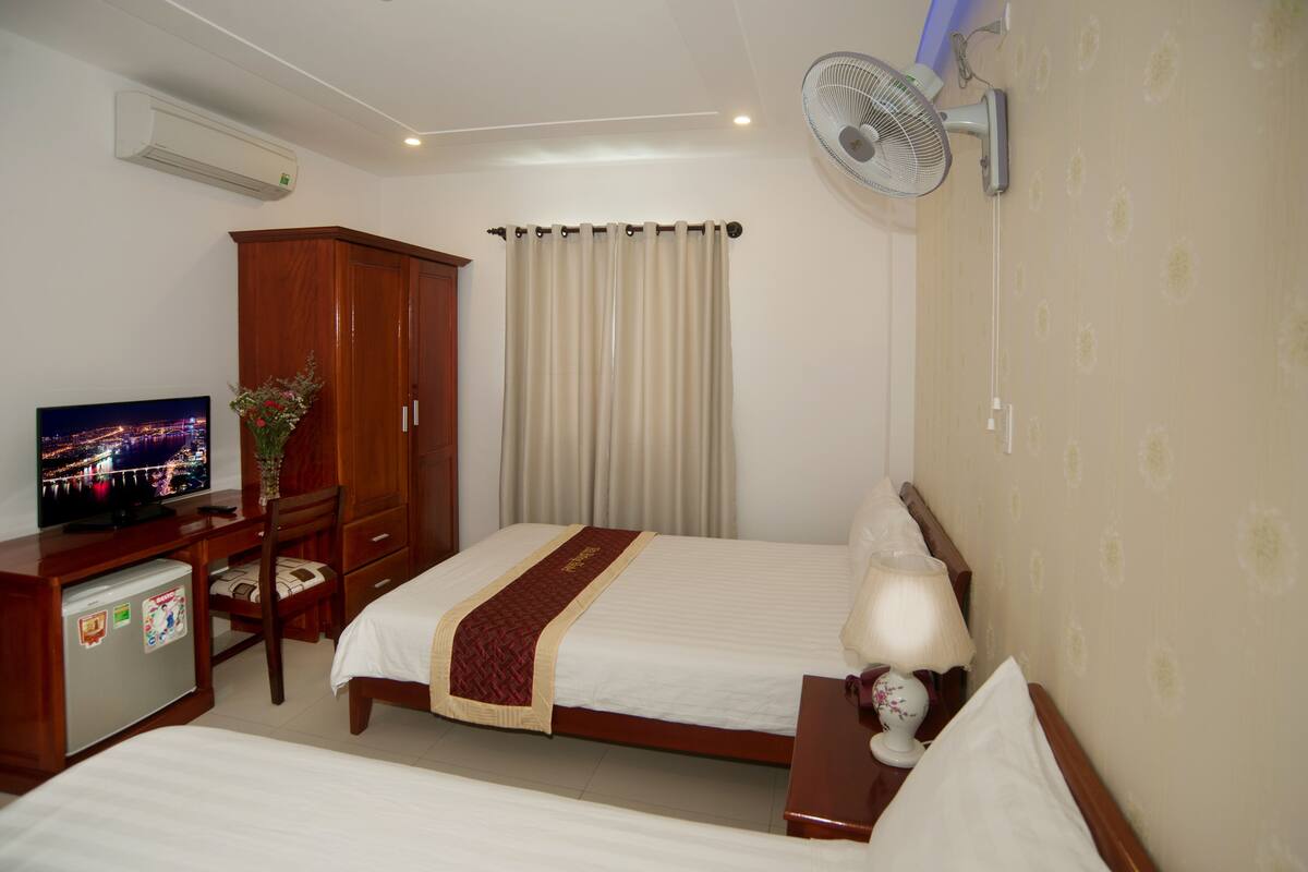 A hotel room with a bed, a desk with a computer, a wardrobe, a wall-mounted fan, and a television displaying a cityscape.