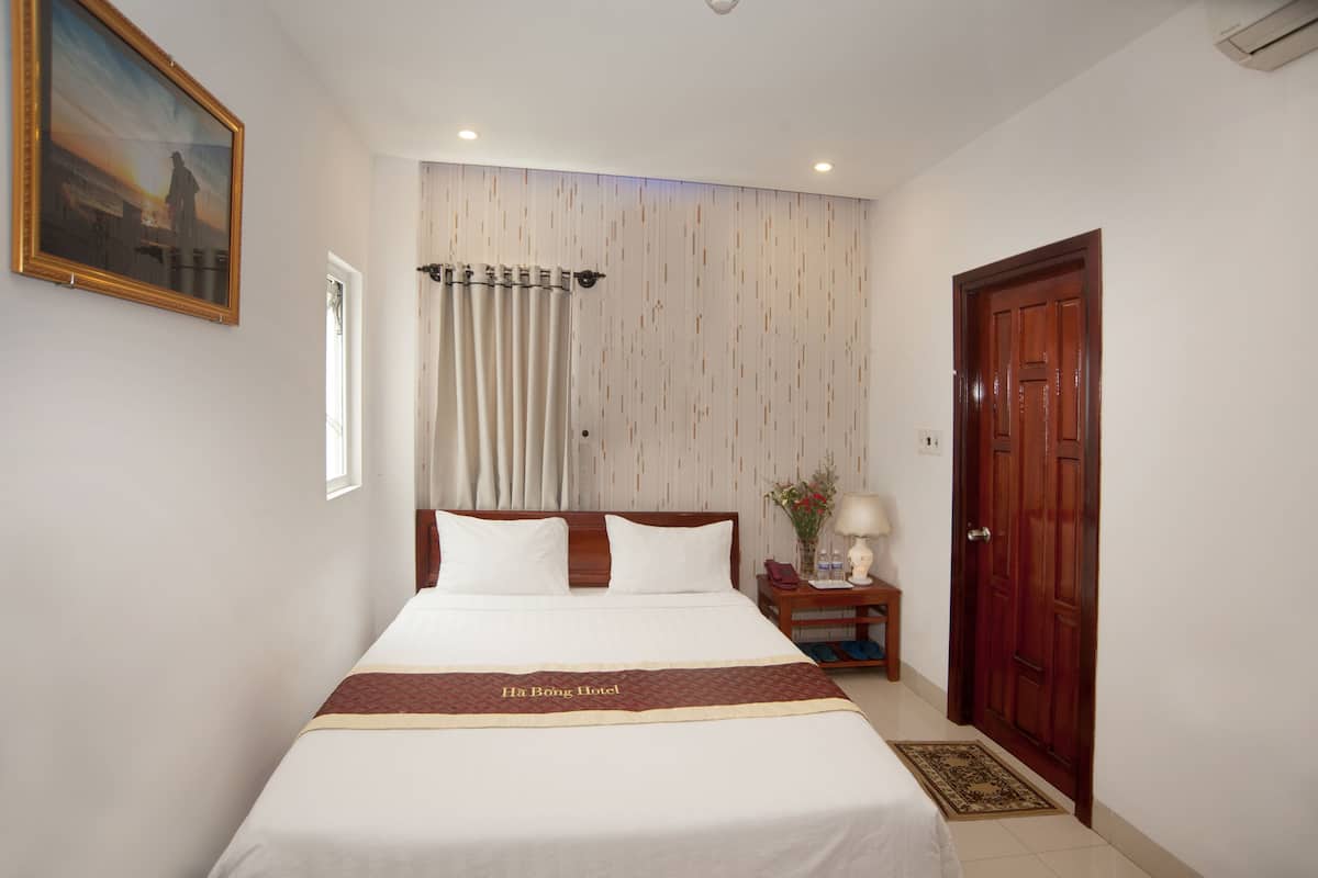 A hotel room with a bed, a nightstand with a lamp, a wooden door, and a painting on the wall.