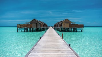 Une jetée en bois menant à des bungalows sur pilotis, au-dessus d’une eau turquoise et cristalline.