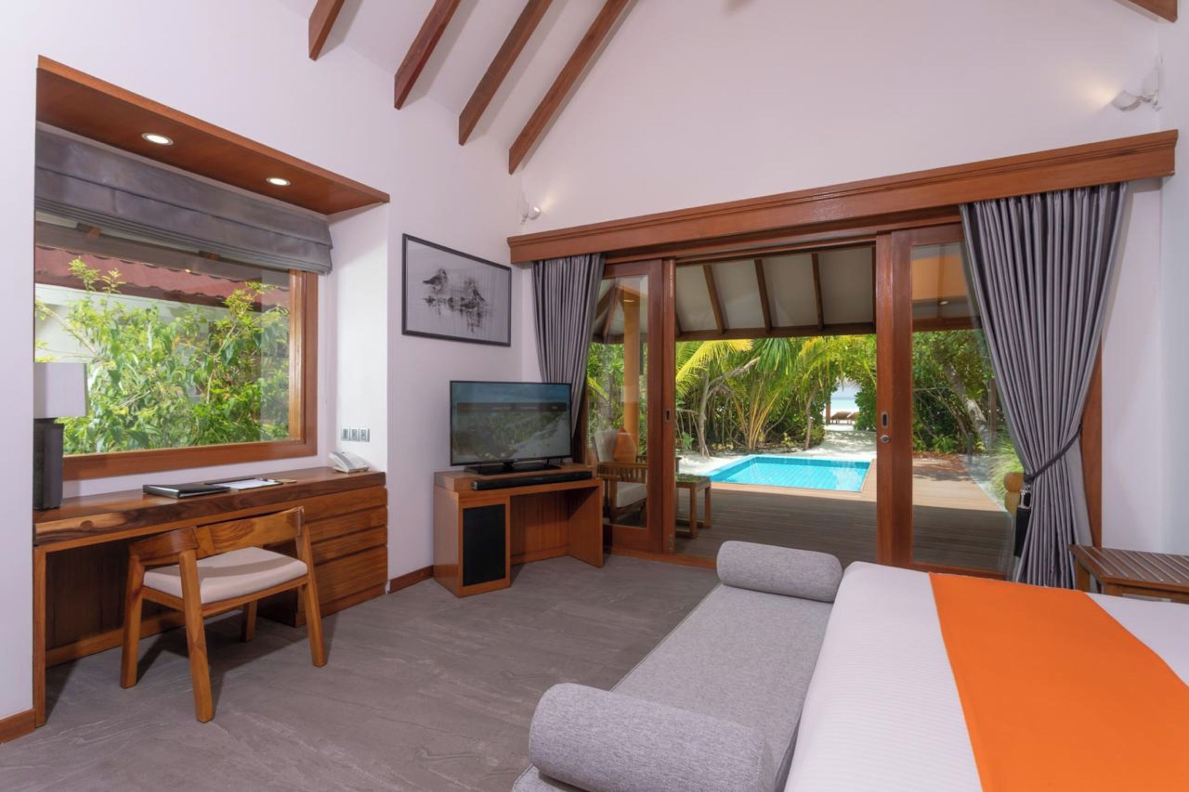 A bedroom with a bed, a desk with a chair, a television, and a view of a swimming pool.