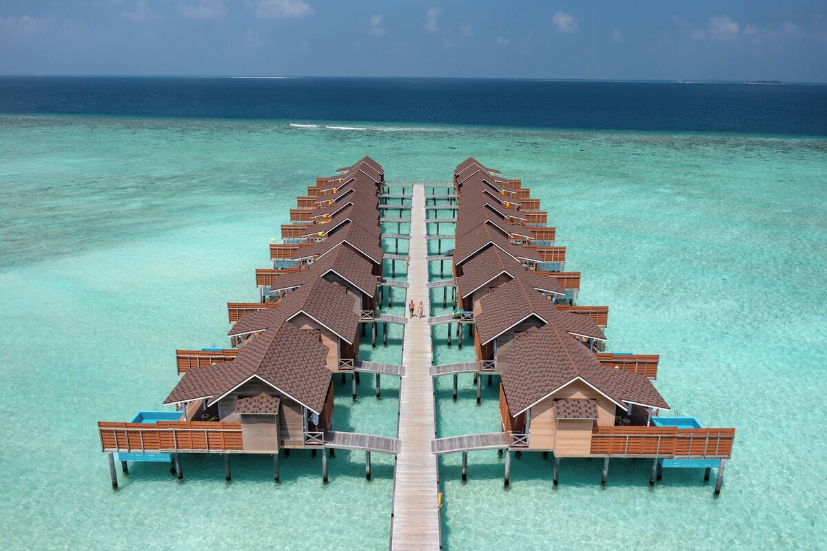 Eine Vogelperspektive zeigt ein Resort mit Bungalows, die über dem Wasser liegen und durch einen zentralen Steg verbunden sind.