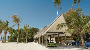 A beach house with a thatched roof, surrounded by palm trees and a sandy beach.