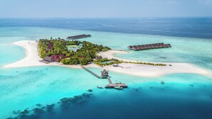 Vista aérea de un resort tropical con bungalows sobre el agua, una playa y aguas turquesas cristalinas.