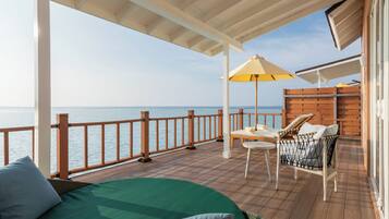 A deck with a view of the sea, a bed, a wicker chair, and a table with an umbrella.