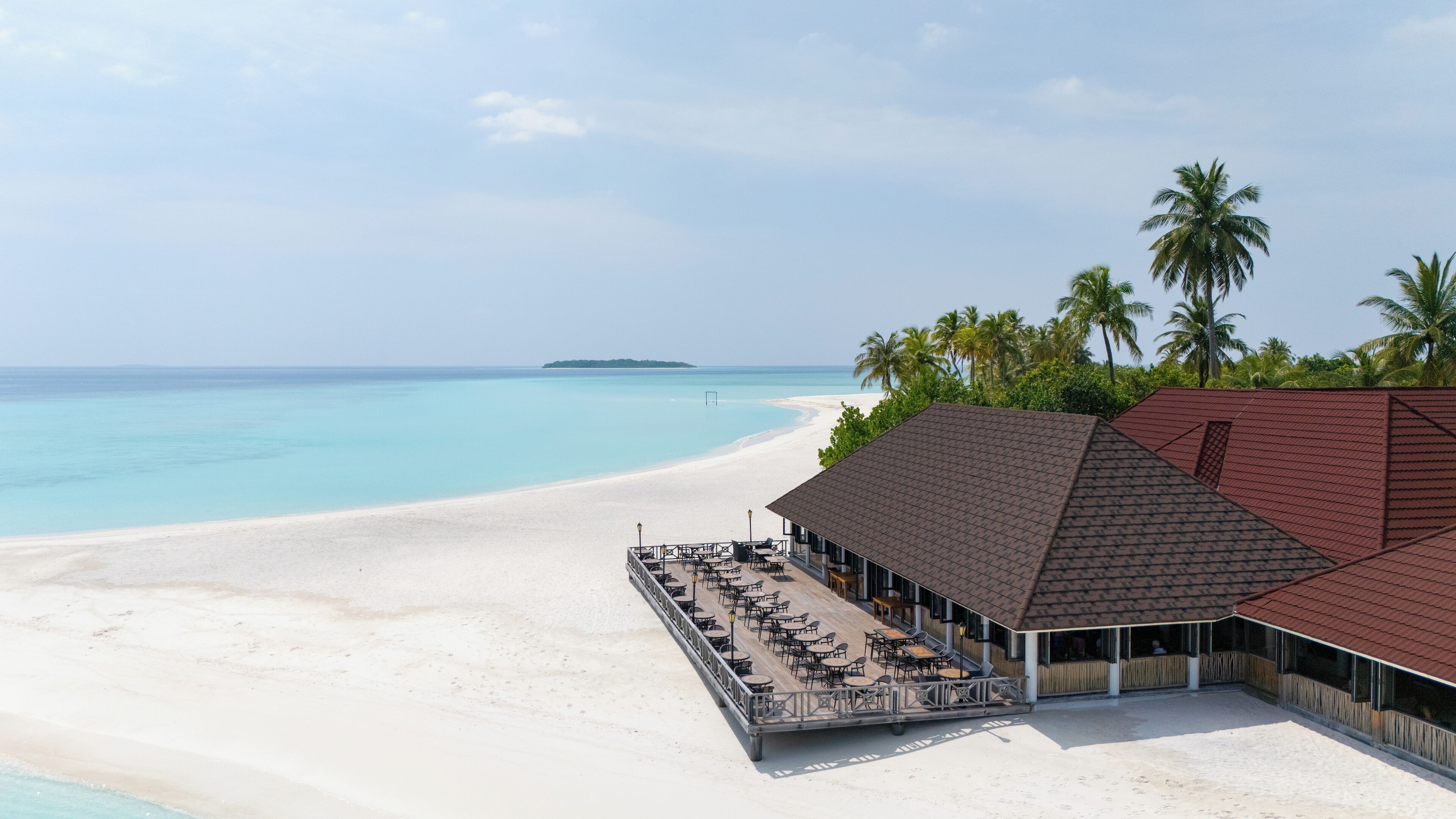 Un hotel frente a la playa con una zona de estar al aire libre cubierta, una playa de arena y palmeras.