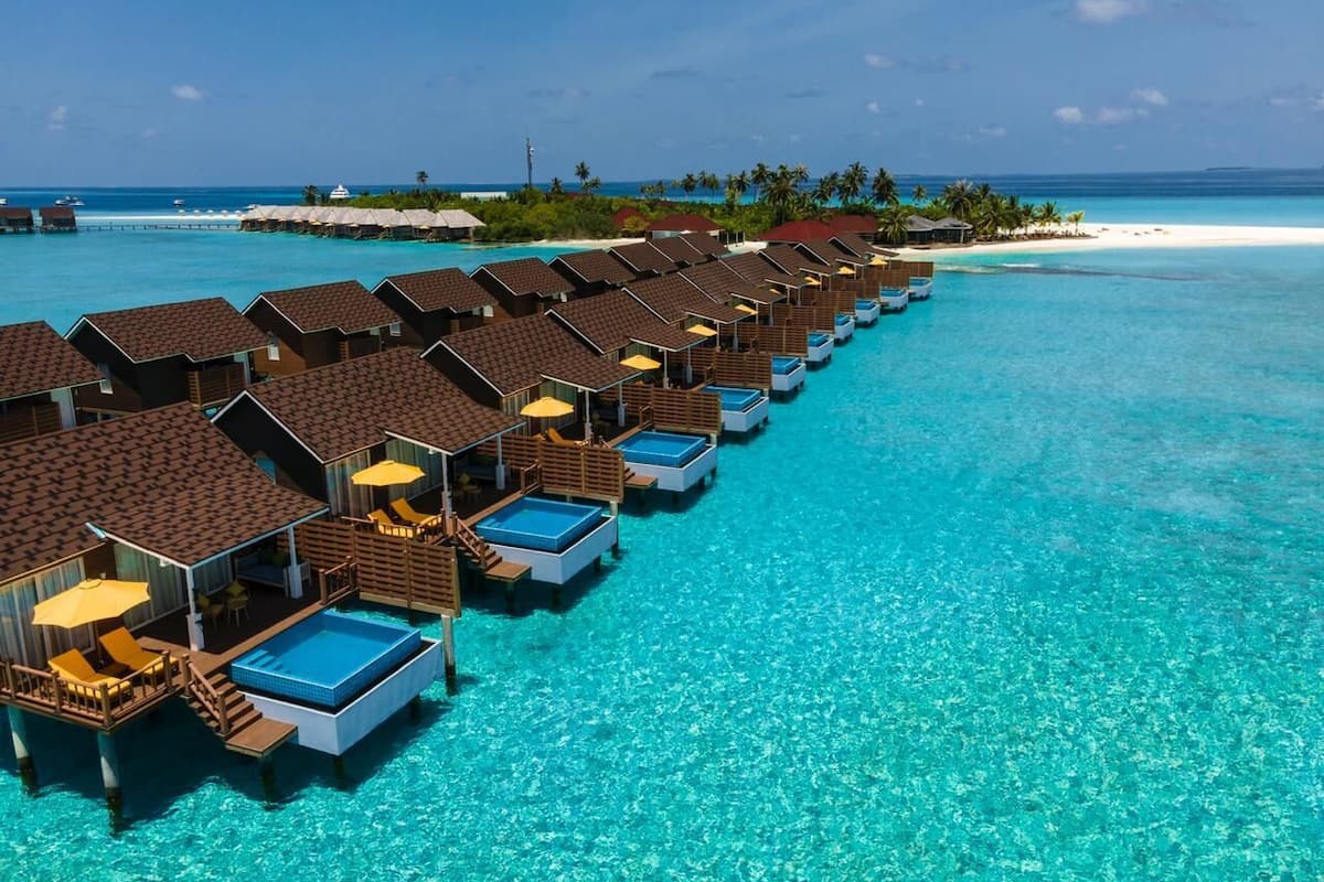 Wasserbungalows mit Privatpools, verbunden durch Wege, mit Blick auf ein klares türkisblaues Meer und einen weißen Sandstrand.