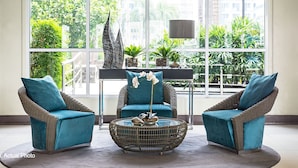 Un espace détente avec des fauteuils en osier et une table basse ronde, de grandes fenêtres offrant une vue sur la verdure et les bâtiments, et une lampe moderne.