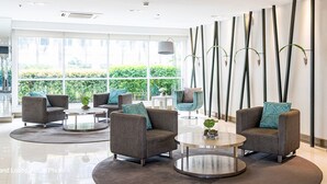 Un hall d’entrée moderne avec trois fauteuils gris, deux tables rondes et une grande fenêtre donnant sur un paysage verdoyant.