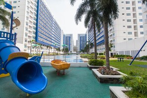 A playground with a blue slide and a yellow bucket, surrounded by palm trees and a pool.