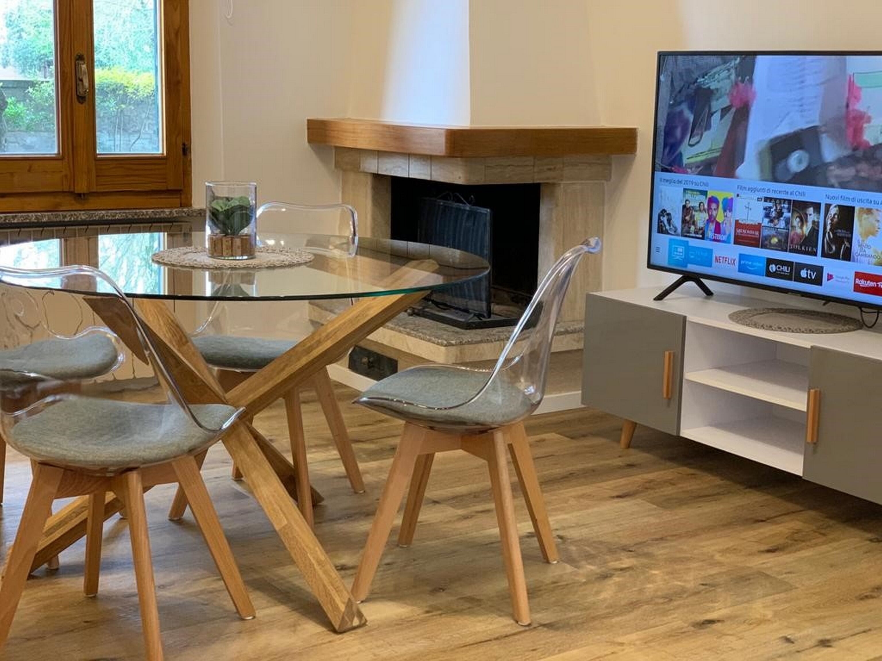 Uma sala de estar moderna com uma mesa de jantar de vidro, cadeiras transparentes, lareira e uma televisão de tela plana montada em um armário.