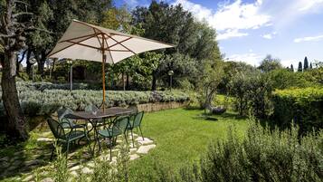 A patio with a table and chairs under a large umbrella, surrounded by lush greenery and a stone pathway.