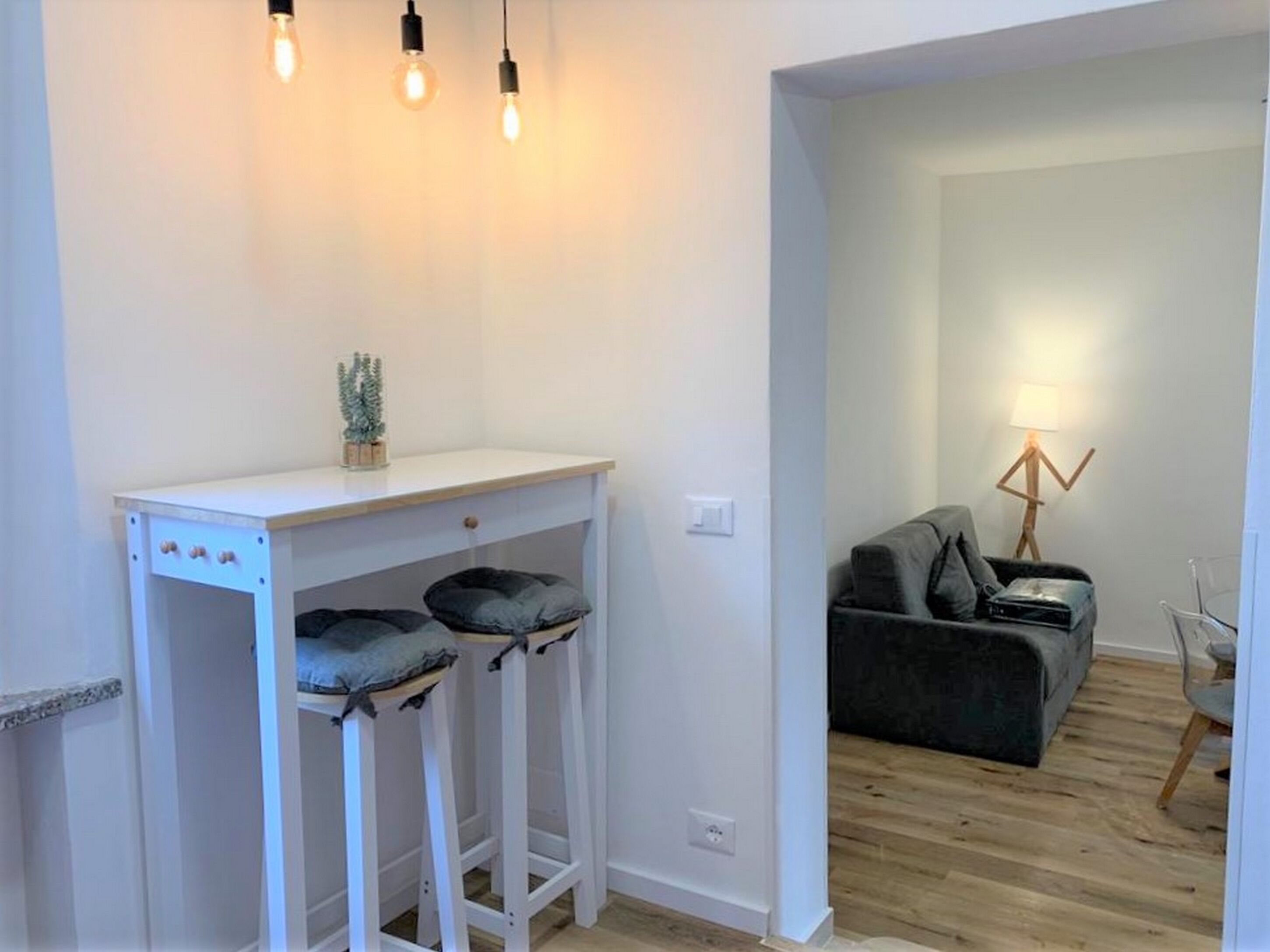 Ein moderner Wohnraum mit einem weißen Schreibtisch, zwei Barhocker, einem grauen Sofa und einer Stehlampe.