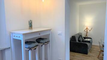 Ein moderner Wohnraum mit einem weißen Schreibtisch, zwei Barhocker, einem grauen Sofa und einer Stehlampe.