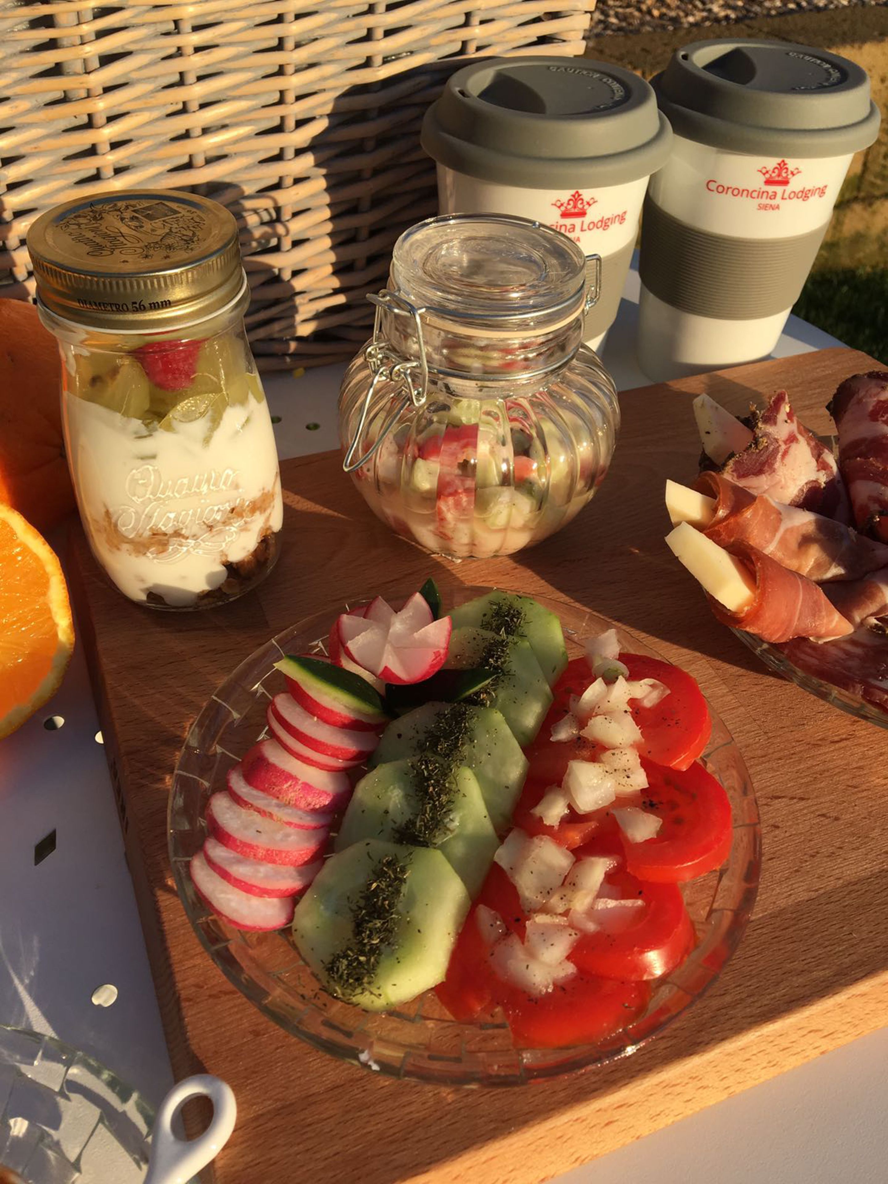 Un pique-nique avec un bocal en verre contenant des aliments disposés en couches, un autre bocal en verre contenant des légumes marinés, et une assiette de légumes et de viande coupés en tranches.