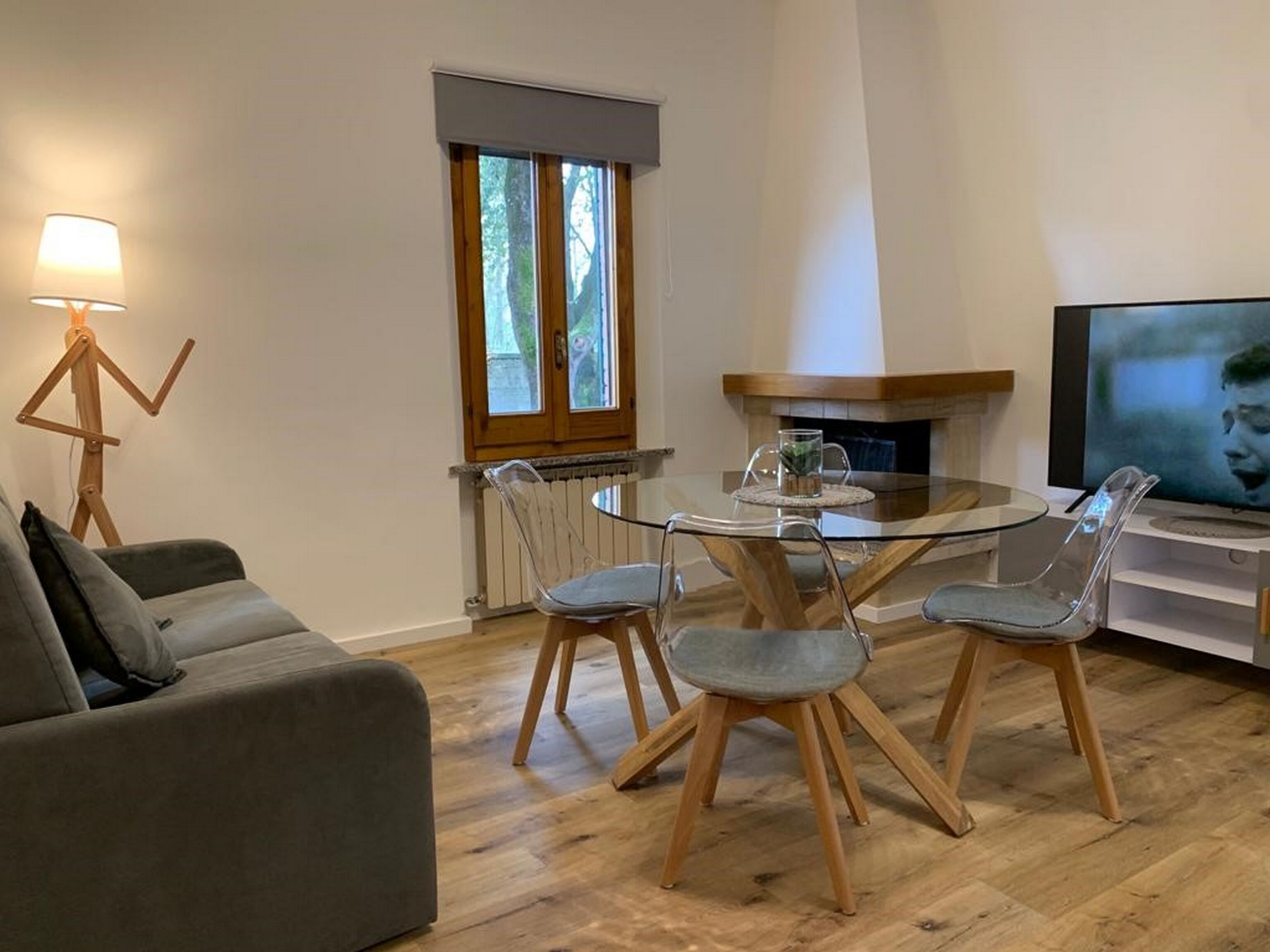Ein modernes Wohnzimmer mit einem Glastisch, transparenten Stühlen, einem grauen Sofa, Holzboden, Kamin und einem Fernseher auf einem Schrank.