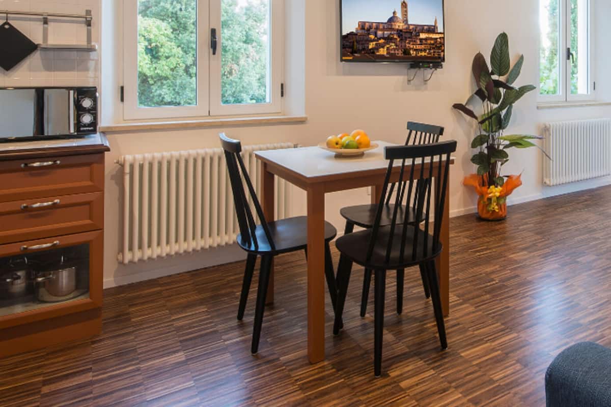 A dining area with a table and chairs, a radiator, a window with a view of a cityscape, and a kitchen area with cabinets and utensils.