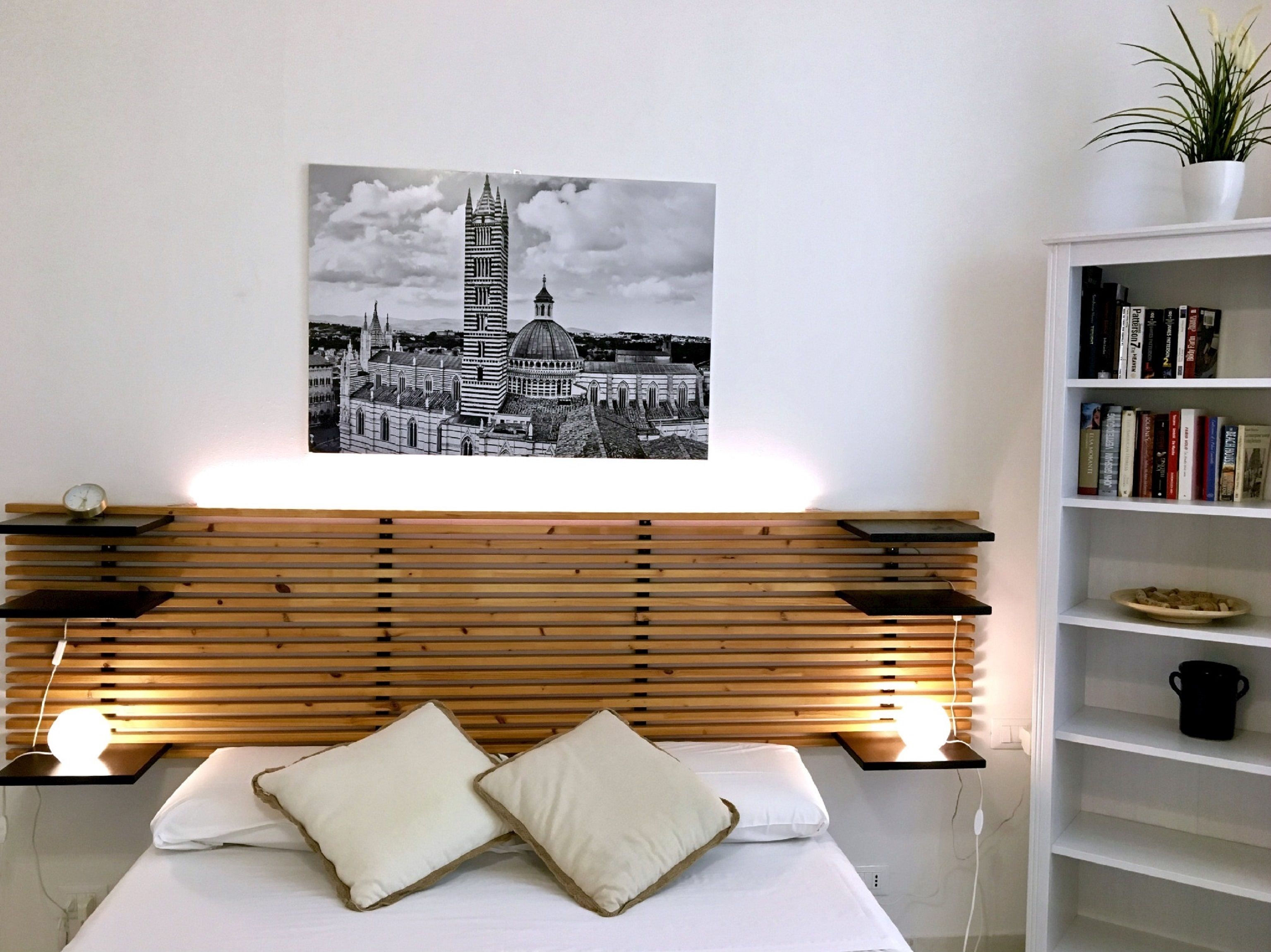 Un dormitorio con cabecera de madera, cama con ropa de cama blanca, dos almohadas, una mesita de noche con un reloj, un cuadro colgado en la pared de un paisaje urbano y una estantería con libros y una planta.