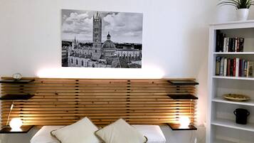 Un dormitorio con cabecera de madera, cama con ropa de cama blanca, dos almohadas, una mesita de noche con un reloj, un cuadro colgado en la pared de un paisaje urbano y una estantería con libros y una planta.