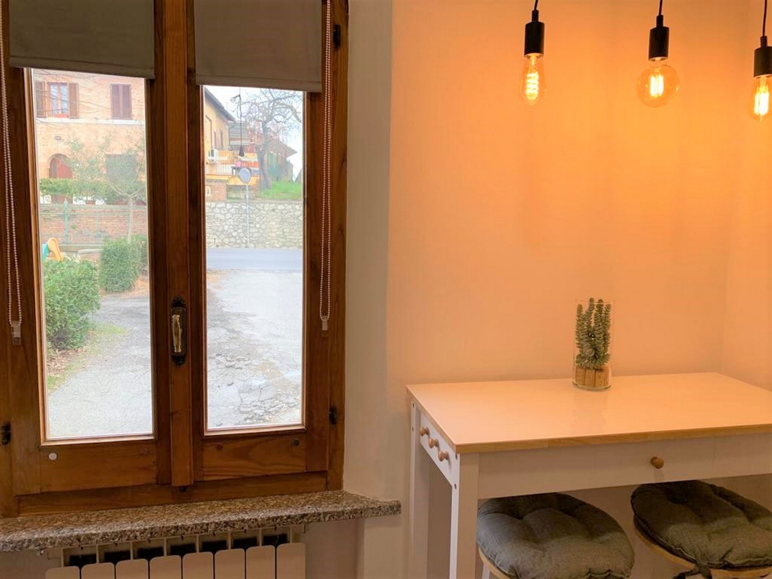 Una habitación con puerta de madera, ventana con vista a un patio, una encimera blanca con una planta y dos asientos con cojines verdes.