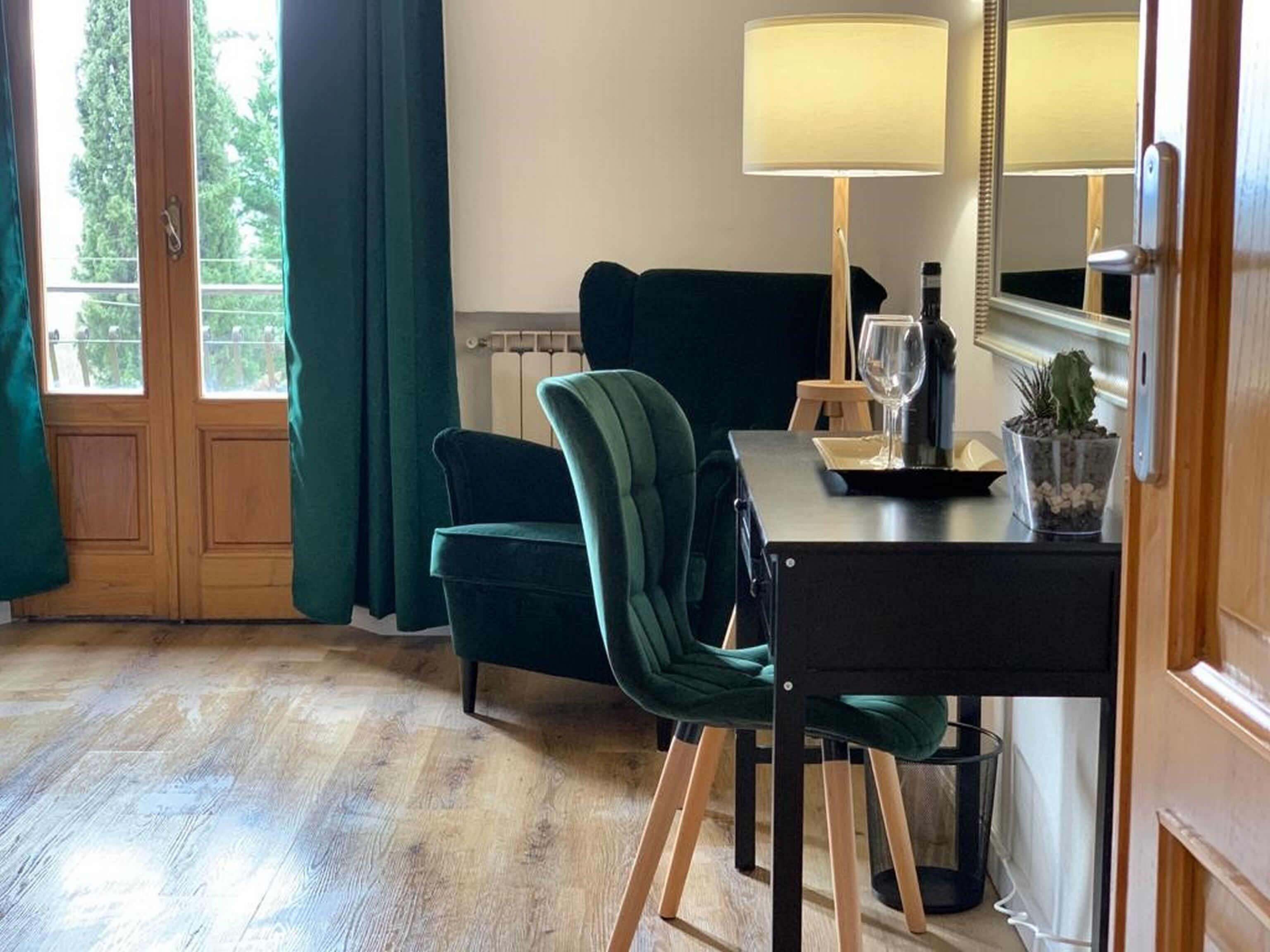 Um quarto com piso de madeira, um sofá verde, uma mesa de jantar com cadeiras, um abajur, uma taça de vinho e uma pequena planta.