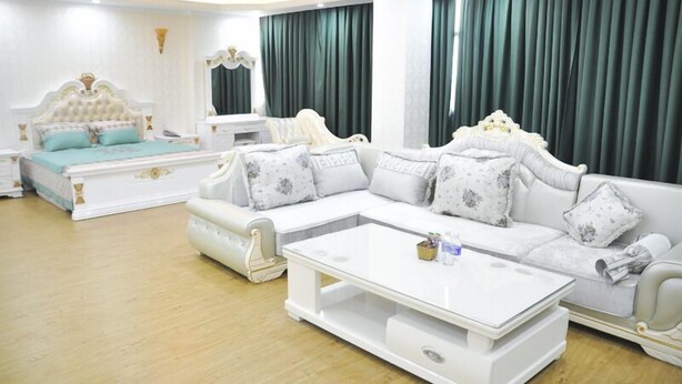 A modern living room with a large white sectional sofa, a matching white coffee table, and a white bed with a tufted headboard. - Phoenix Hotel (Hac Thanh)