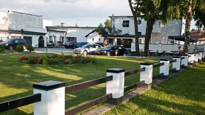 A parking lot with several cars, a grassy area with flowers, and a row of white buildings with black railings. - Milano Garden - City Bungalow Hotell, i gränssnittet till Kullabygdens pärlor (Astorp)