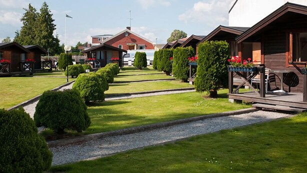A row of wooden cabins with small front porches, neatly trimmed lawns, and a gravel path leading through the property. - Milano Garden - City Bungalow Hotell, i gränssnittet till Kullabygdens pärlor (Astorp)