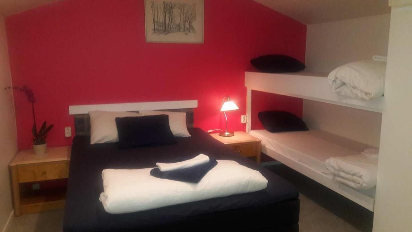A room with a large bed, a bedside table with a lamp, a lower bunk bed, and a red wall with a framed picture.