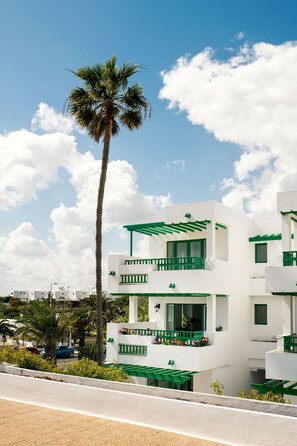 A modern white building with green accents, featuring balconies and a palm tree in the background. - Apartamentos Nazaret (Teguise)