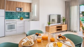 A modern kitchen with white cabinets, a microwave, and a sink. There is a dining table with breakfast items, including croissants, orange juice, and a pitcher. A television is on a wooden stand, and there are plants on the windowsill. - Apartamentos Nazaret (Teguise)