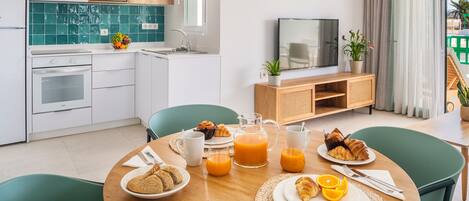Una cocina moderna con armarios blancos, un microondas y un lavabo. Hay una mesa de comedor con alimentos para el desayuno, incluyendo cruasanes, zumo de naranja y una jarra. Un televisor está sobre un soporte de madera, y hay plantas en el alféizar.