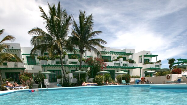A clear swimming pool with a view of a resort building in the background. - Apartamentos Nazaret (Teguise)