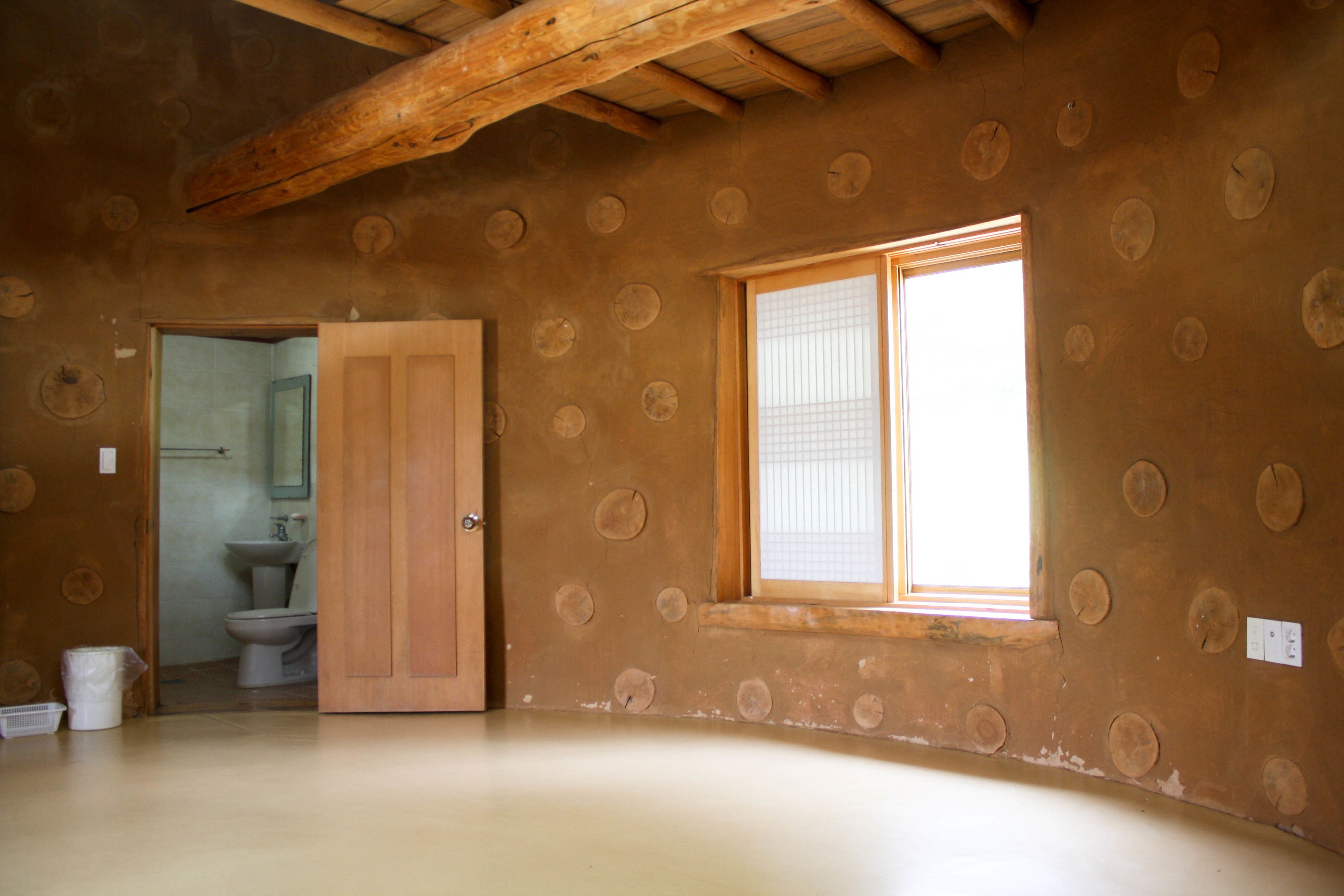 A room with exposed wooden beams, a wooden door, a window with blinds, and a bathroom with a toilet and sink.
