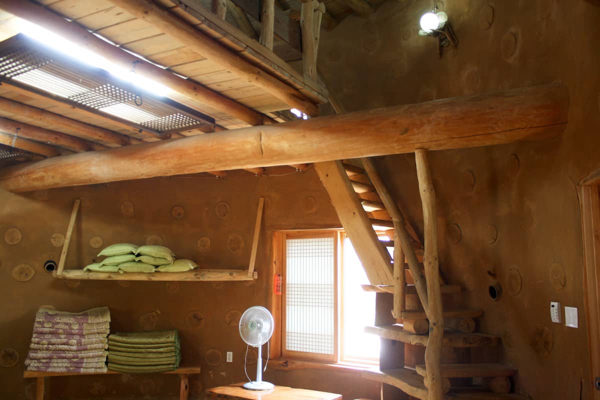 Een rustiek houten interieur met een ventilator, planken met opgevouwen dekens, en een raam met een ruitpatroon.