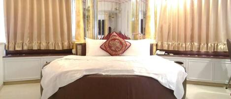 A large, round bed with a white and brown color scheme, positioned in a room with beige curtains and a large mirror.