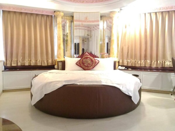 A large, round bed with a white and brown color scheme, positioned in a room with beige curtains and a large mirror.