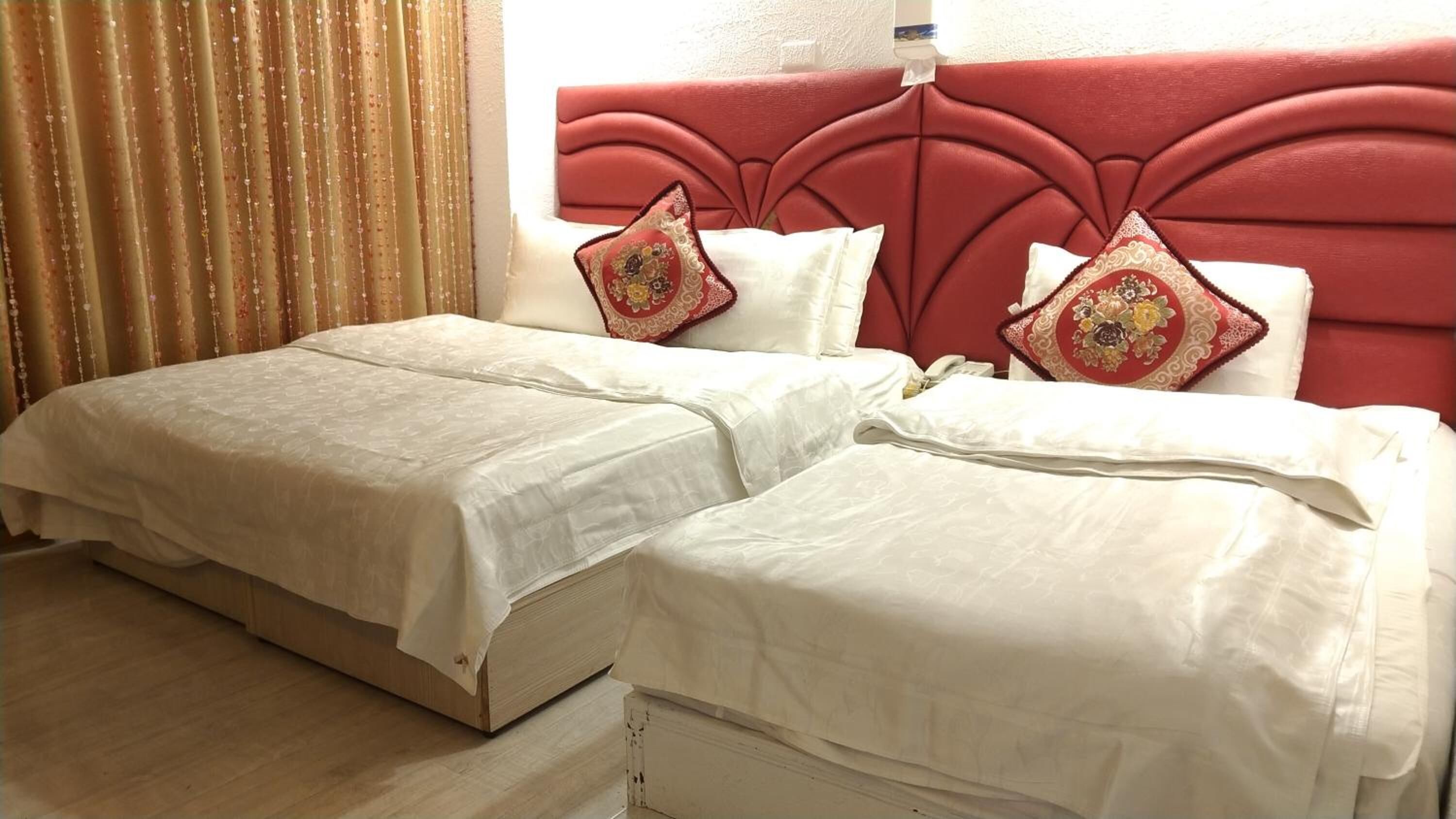 A double bed with white linens and red headboard, two pillows, and a curtain with a floral pattern.