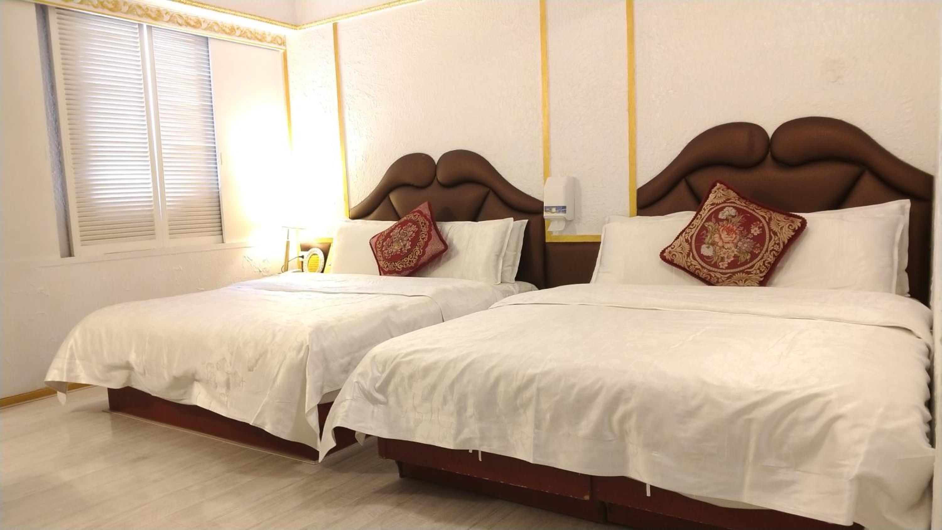 Habitación de hotel con dos camas, cada una con ropa blanca y almohadones decorativos. El piso es de madera, tiene una ventana con persianas y una mesita de noche con una lámpara.