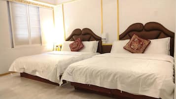 Habitación de hotel con dos camas, cada una con ropa blanca y almohadones decorativos. El piso es de madera, tiene una ventana con persianas y una mesita de noche con una lámpara.