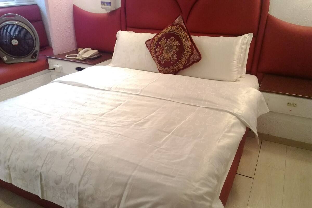 Habitación de hotel con una cama, cabecera roja, cubrecama blanco, almohada decorativa, mesita de noche con un ventilador y un teléfono.