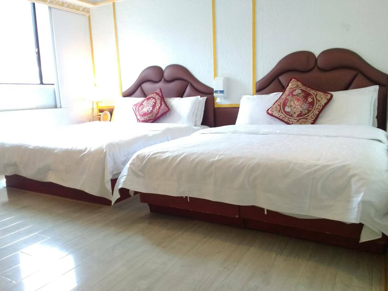 Dos camas dobles con sábanas blancas y almohadas decorativas, suelo de madera y una ventana con cortinas.