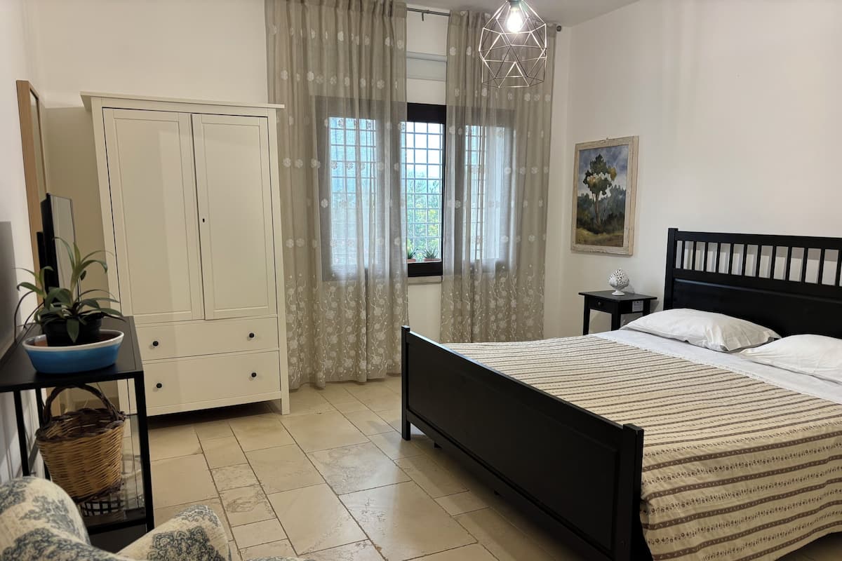 Un dormitorio con una cama, un armario, una silla, una mesita con una planta y un cuadro en la pared.