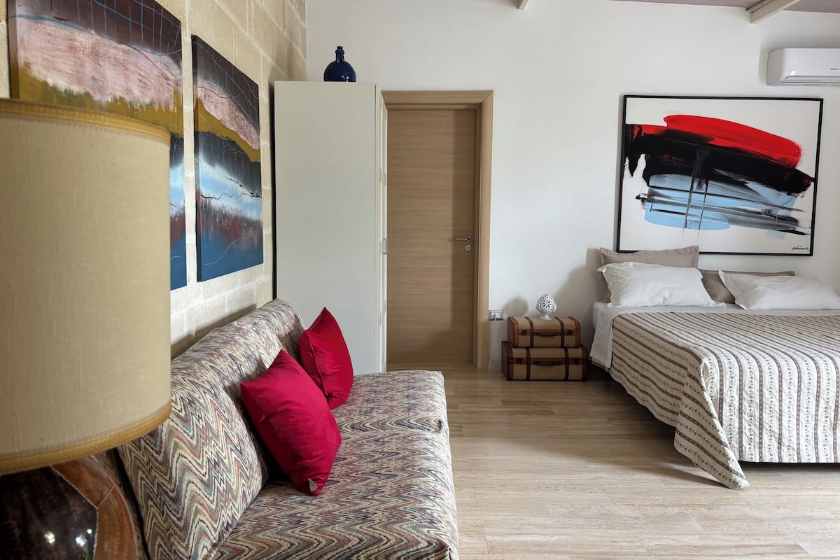 Un dormitorio con un sofá estampado, una cama con ropa de cama a rayas, un cuadro abstracto grande y una maleta en el suelo.