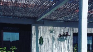 A covered outdoor seating area with a chair and a table, a wall-mounted planter with flowers, and a door with a curtain.