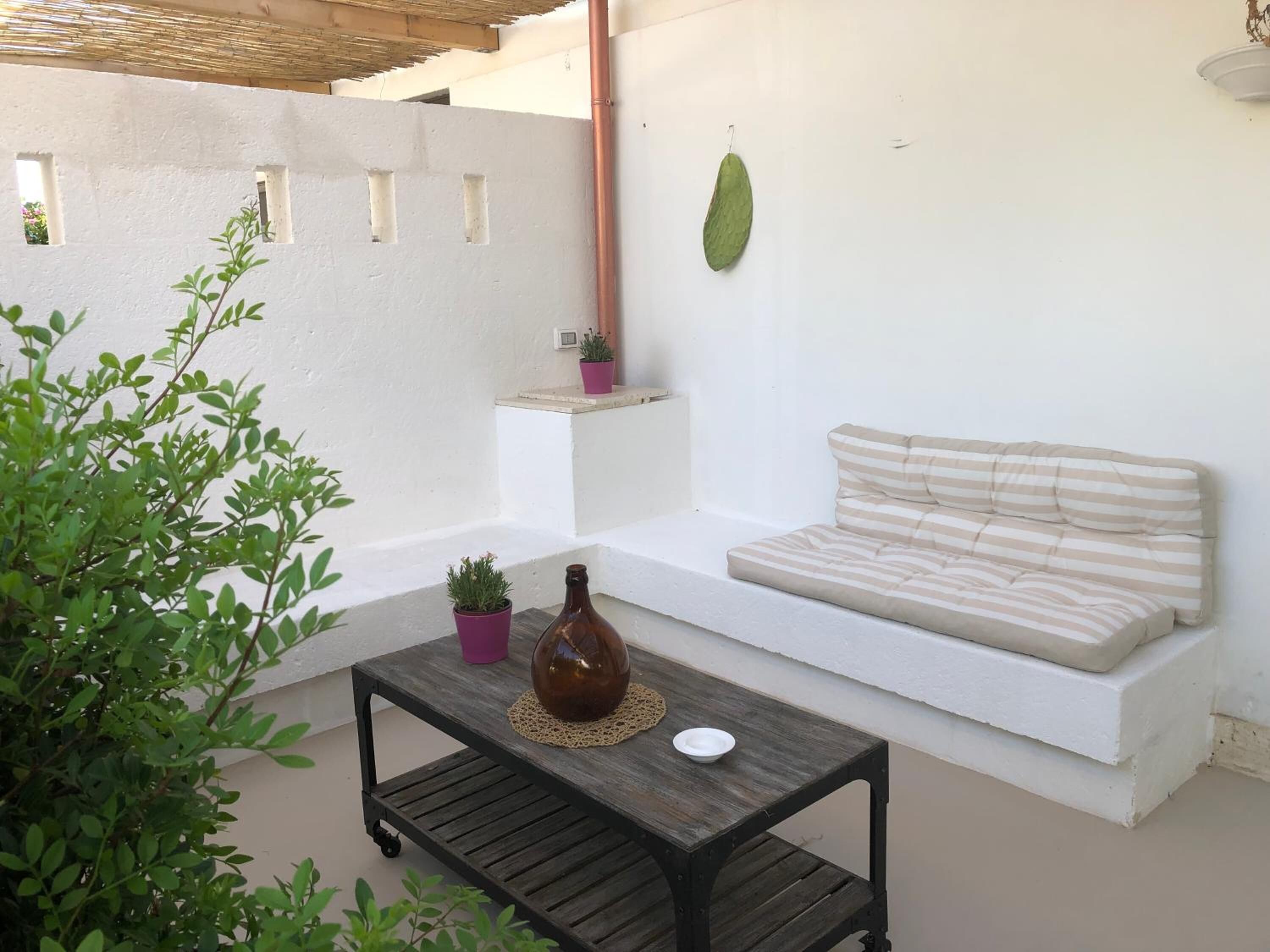 A small outdoor seating area with a cushioned bench and a table, surrounded by white walls and potted plants.