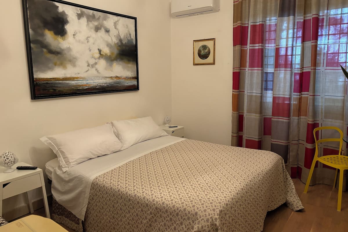 Un dormitorio con una cama, una mesita de noche, una lámpara, un cuadro enmarcado y una ventana con cortinas.