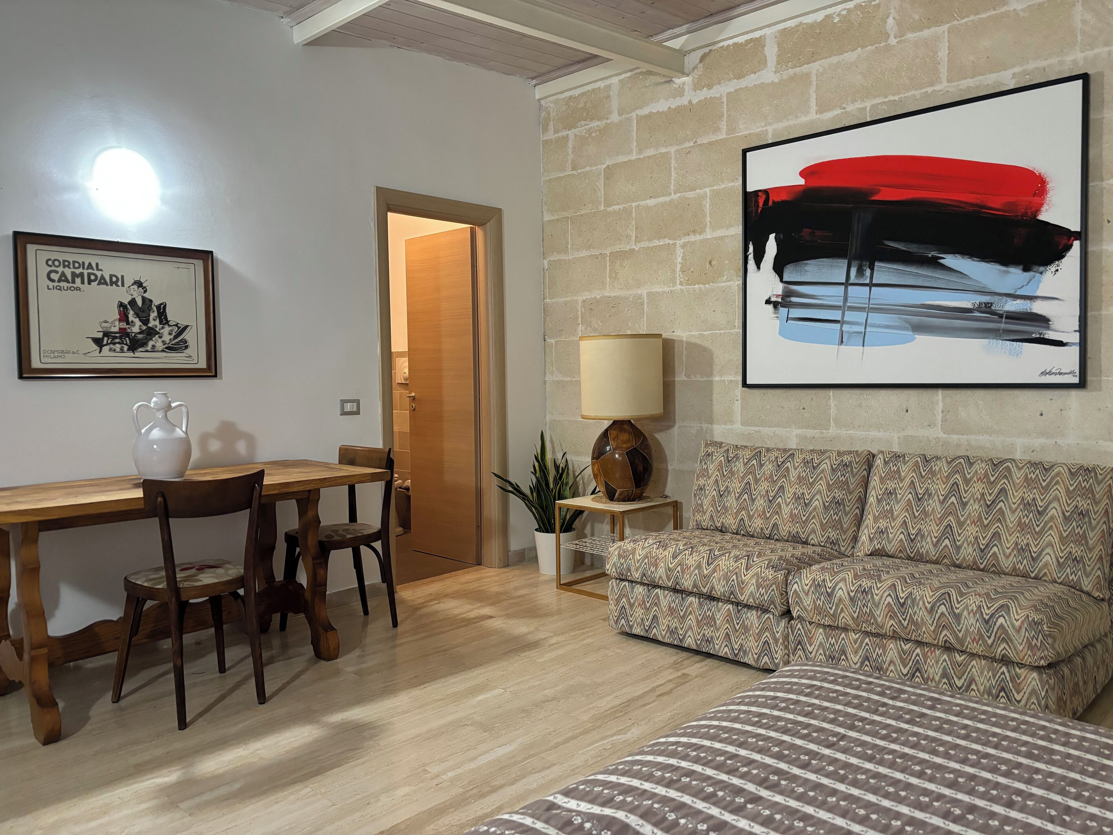 A living room with a wooden dining table, two chairs, a patterned sofa, a lamp, and a painting on the wall.