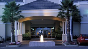 A hotel entrance with a fountain, palm trees, and a red car parked outside.