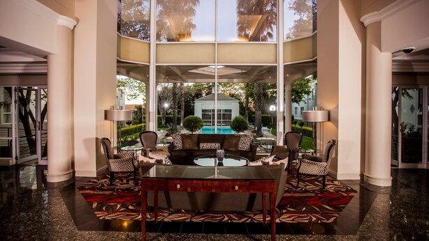 A spacious lobby with a central glass table surrounded by chairs, a pool visible through large windows, and a well-lit interior.
