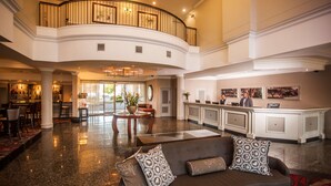 A spacious lobby with a curved reception desk, a seating area with a sofa and armchairs, and a high ceiling with a balcony.